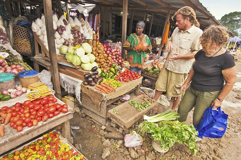 Two Western travellers buy vegetables at a market stall.