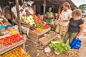 Two Western travellers buy vegetables at a market stall.