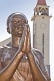 Bronze painted statue of woman praying in front of the white stucco Roman Catholic church.
