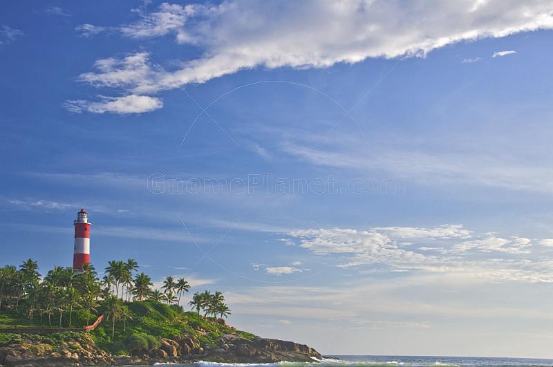 Red and white bands of Vizhinjam Lighthouse tower, set amongst coconut palm trees and a wide open blue sky.