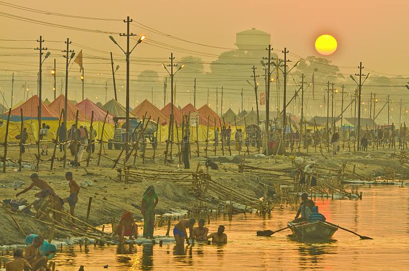 Rowing boat passes bathing pilgrims and tents next to River Ganges in early dawn light.