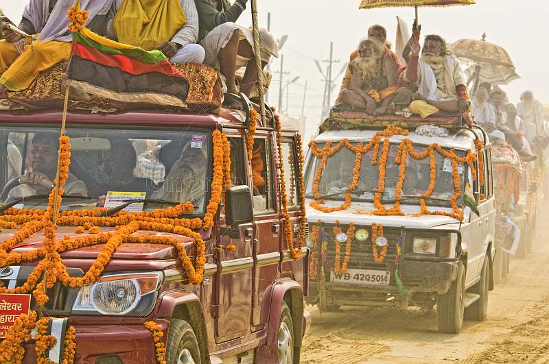 Decorated trucks drive through dust clouds in Basant Panchami Snana procession.
