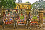 Three colorful decorated bicycle rickshaws next to old city wall.