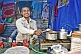 Image of Smiling tea-stall owner adds sugar to pan of boiling Indian chai tea.