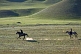 Image of Two Kyrgyz horsemen riding at a canter over sparse grassland.