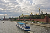 A sight-seeing boat on the Moscow River passes the red walls of the Kremlin, under a cloudy sky.