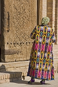 Elderly Uzbek lady in traditional hand dyed silk Chopon coat, stands before an intricately carved wooden door.
