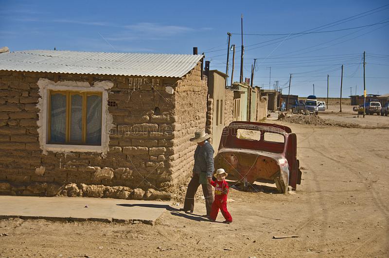 Woman and child walk past shell of car on street of adobe houses.