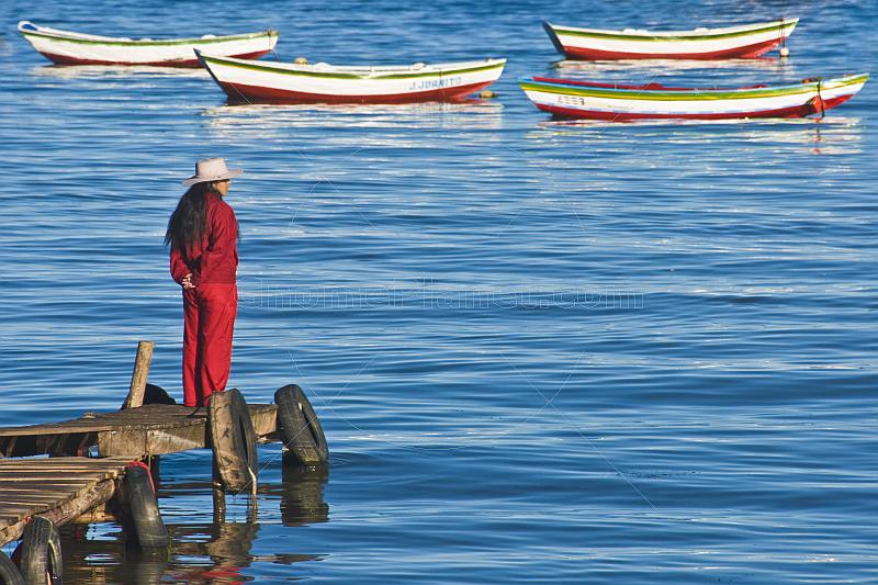 Woman in hat and red coat and trousers looks at moored rowing boats.