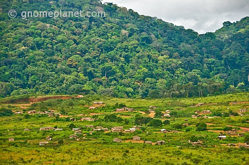 A village of wooden huts occupies cleared rain forest land below tree-covered hills.