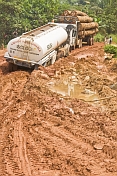 A timber truck tows a fuel tanker through a muddy and deeply rutted section of logging road.