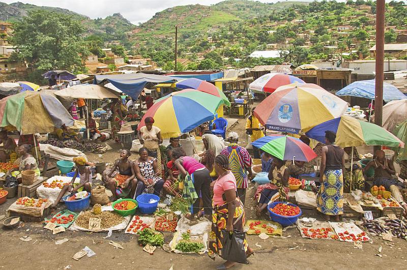 Women under umbrellas selling vegetables in a crowded market with views of hillside to rear.