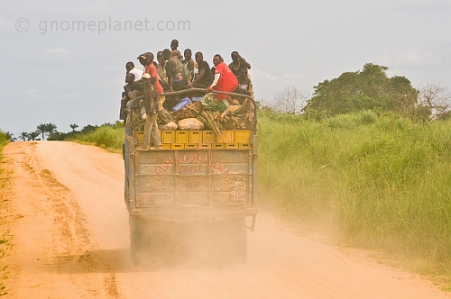 A heavily laden truck with passengers riding on top of the cargo drives down a dusty main road.