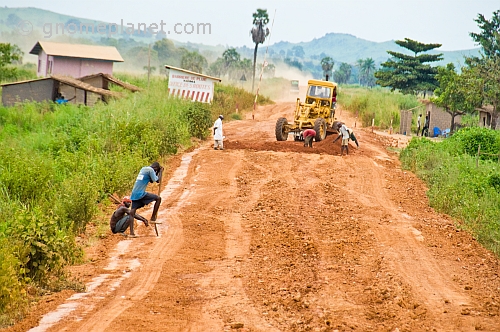 A grader repairs a rough and rocky road whilst workmen rest on their shovels.