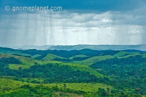 Rain falls from dark clouds over the hills and valleys of forested countryside.