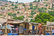 People pass by foodstalls whilst in the background is a hillside covered with brick houses and shacks.