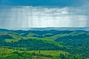 Rain falls from dark clouds over the hills and valleys of forested countryside.