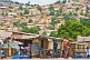 Image of People pass by foodstalls whilst in the background is a hillside covered with brick houses and shacks.